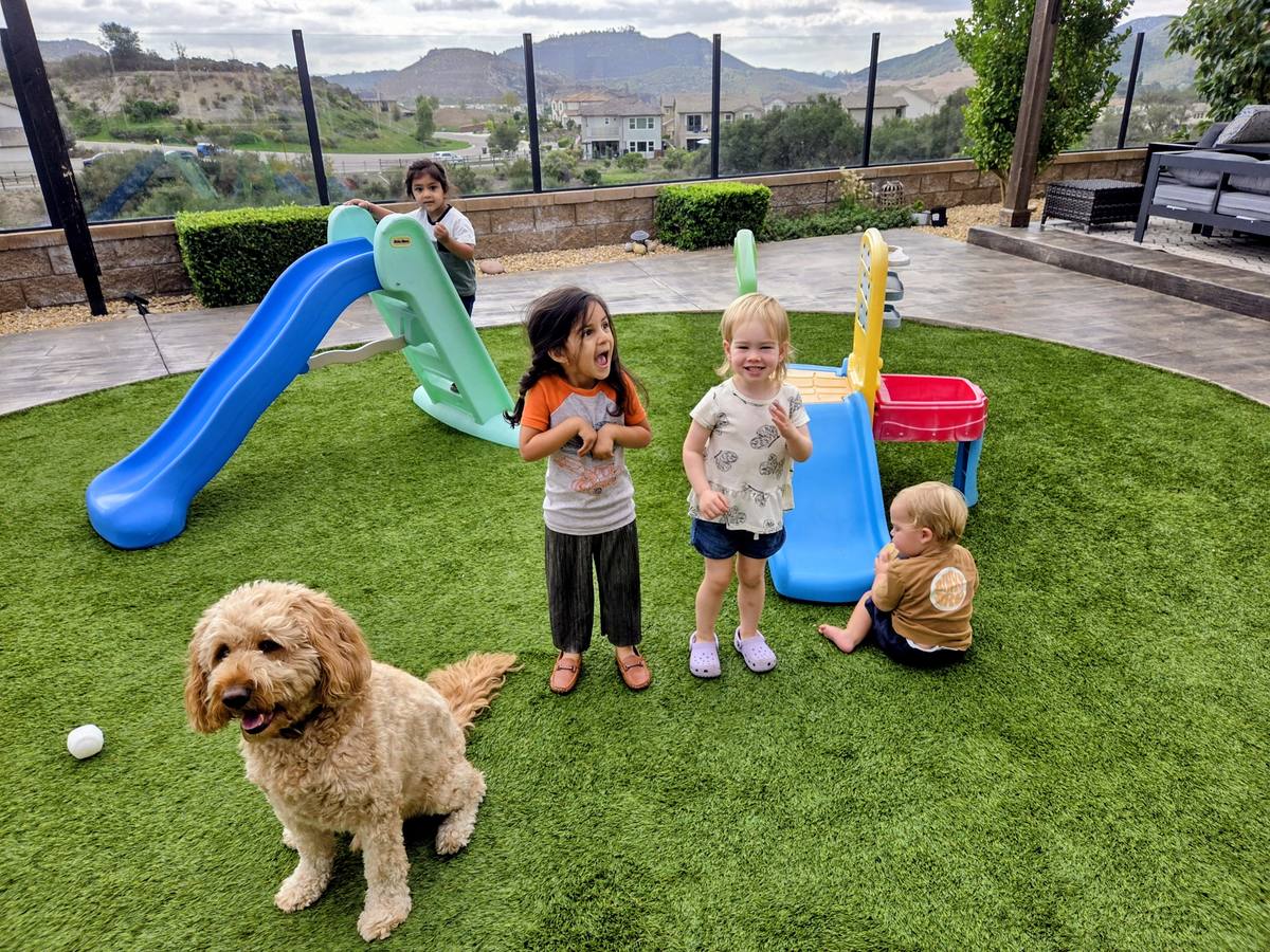 Toddlers playing with a soccer ball and climbing frame on the artificial turf, with a covered patio and hills in the background.