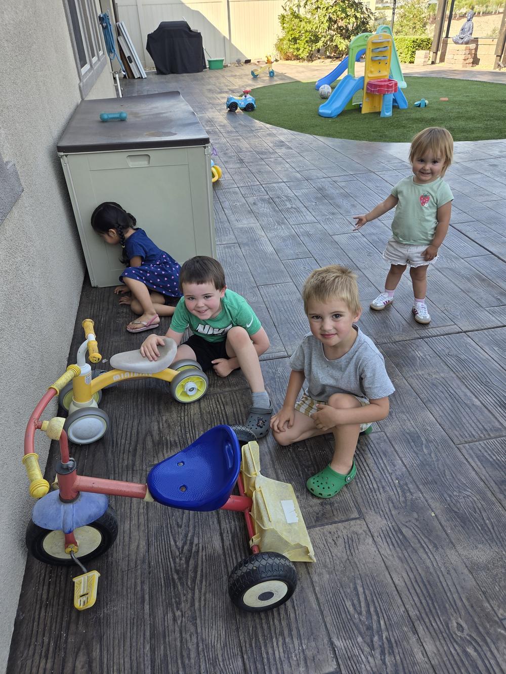 Children sitting on the shaded patio with trikes and ride-on toys, next to the turf play area.