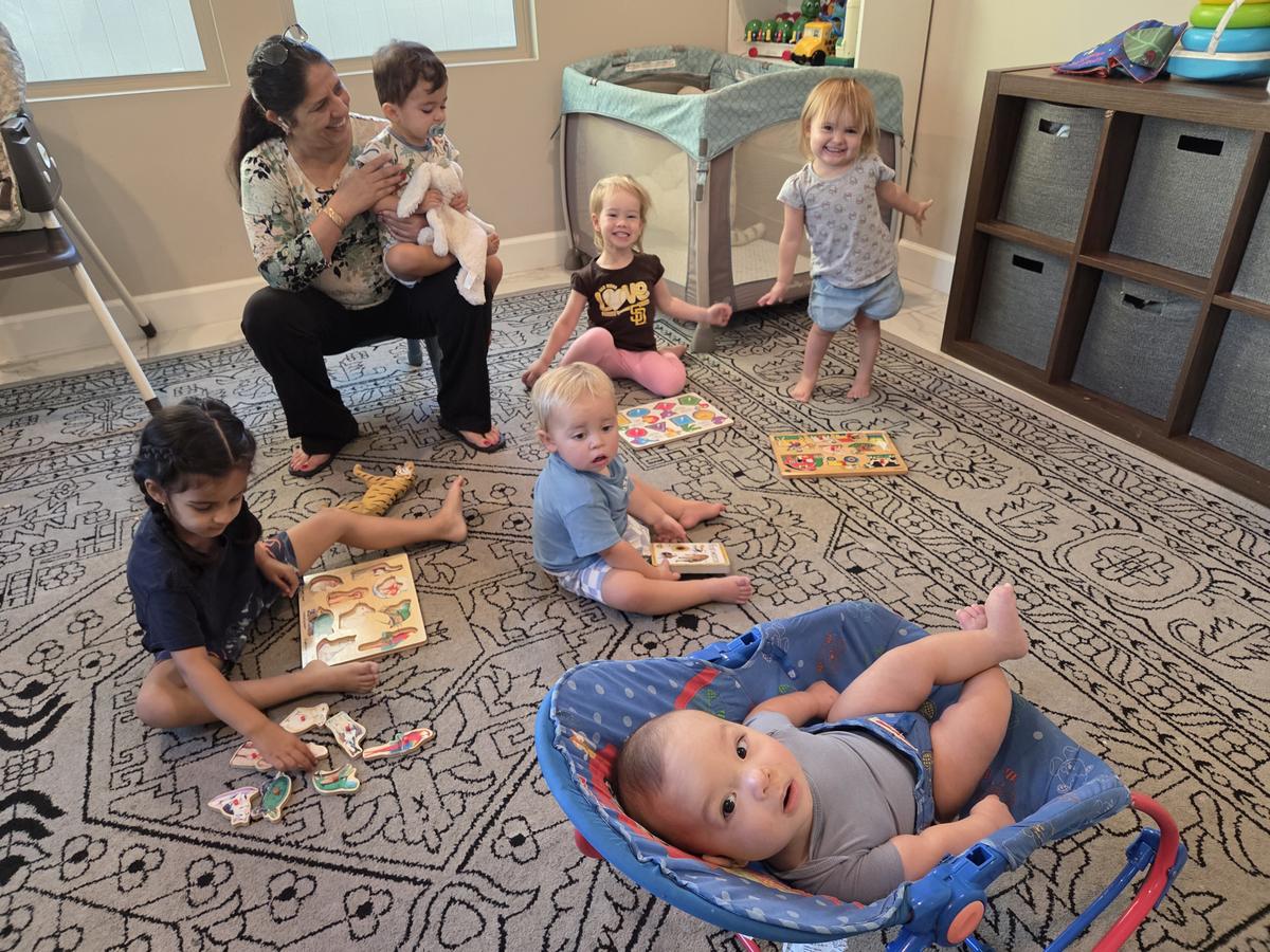 Children working on wooden puzzles on the playroom rug while a caregiver holds a toddler nearby.
