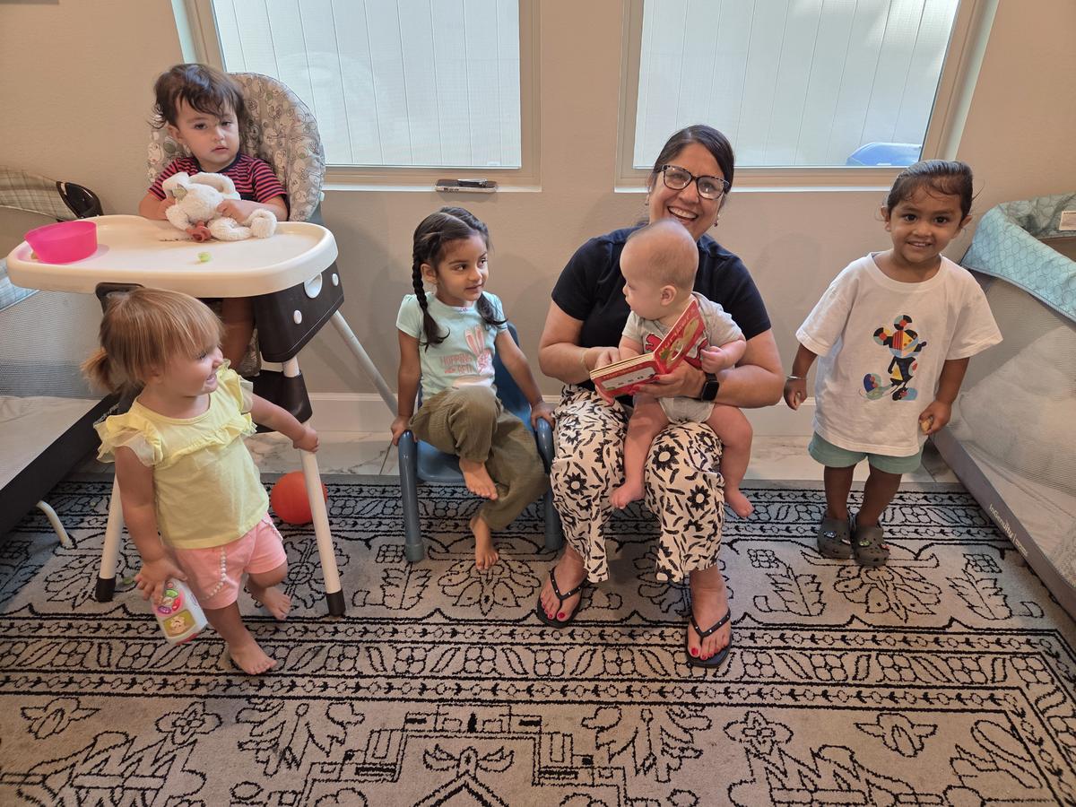 A caregiver sits with a group of children in the playroom, holding a baby and reading a numbers board book aloud.