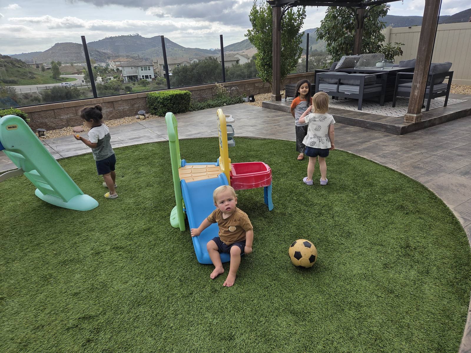 Four children playing on a slide and climber in the fenced backyard, with the Harmony Grove hills behind them and the daycare's dog on the grass.